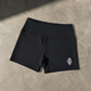 Black athletic shorts with a wide waistband and a small white stylized logo on the lower left leg, laid flat on a textured gray concrete surface.
