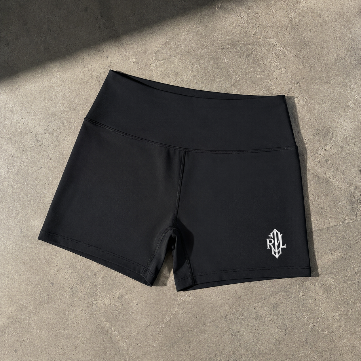 Black athletic shorts with a wide waistband and a small white stylized logo on the lower left leg, laid flat on a textured gray concrete surface.