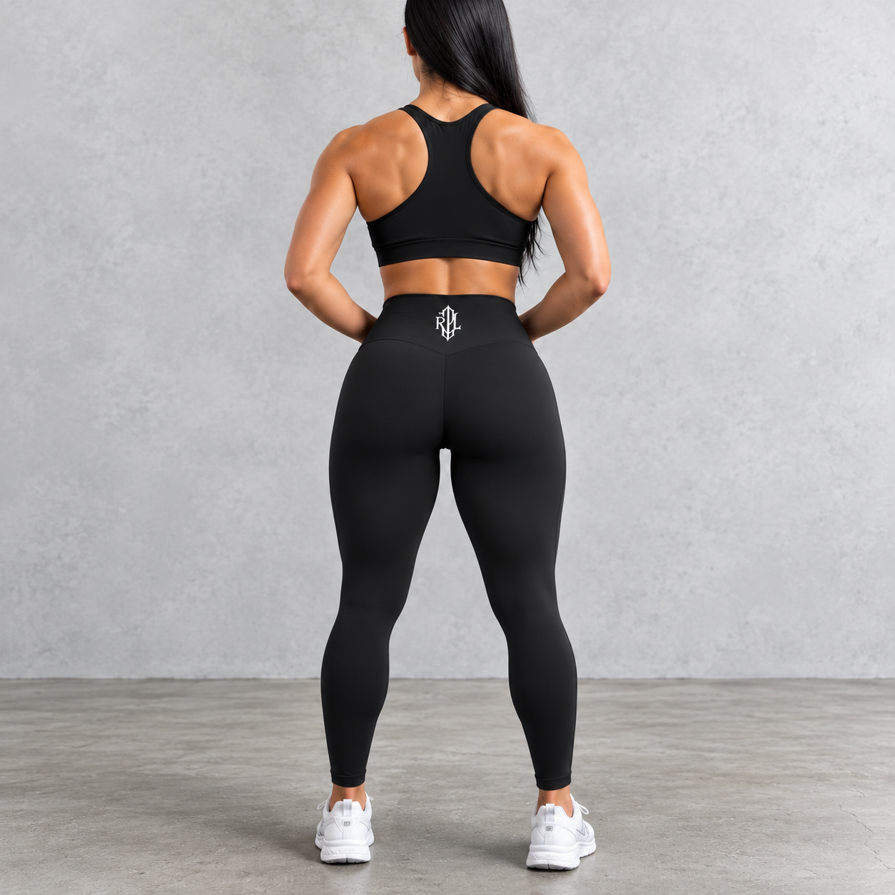 Rear view of a woman wearing a black athletic sports bra and high-waisted black leggings with a small white geometric logo on the back waistband, standing against a textured gray studio wall.