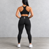 Rear view of a woman wearing a black athletic sports bra and high-waisted black leggings with a small white geometric logo on the back waistband, standing against a textured gray studio wall.