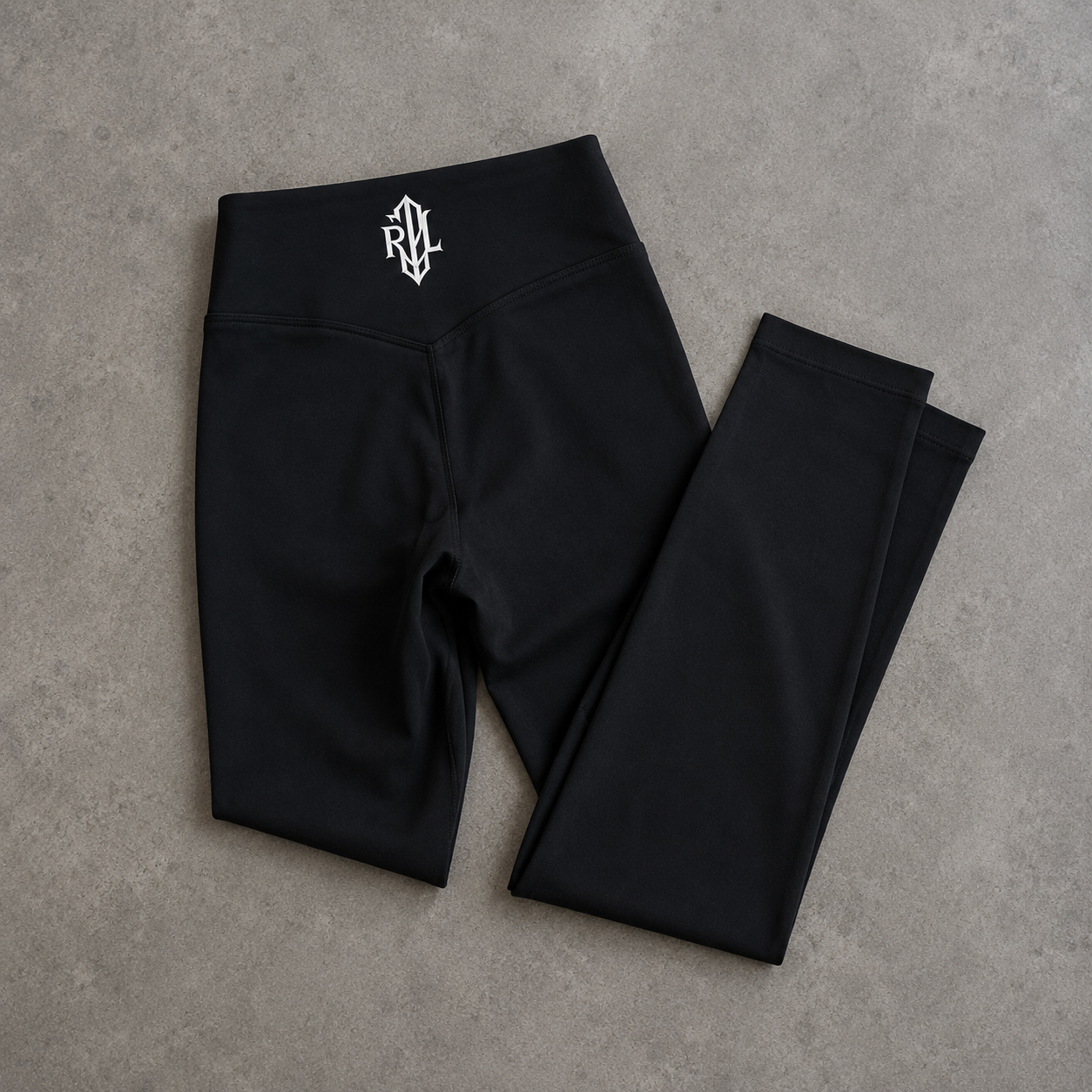 Black high-waisted leggings with a white stylized 'RVL' logo on the waistband, laid flat on a textured gray concrete surface.