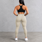 Rear view of a woman wearing beige high-waisted leggings and a black sports bra against a gray concrete wall and floor.
