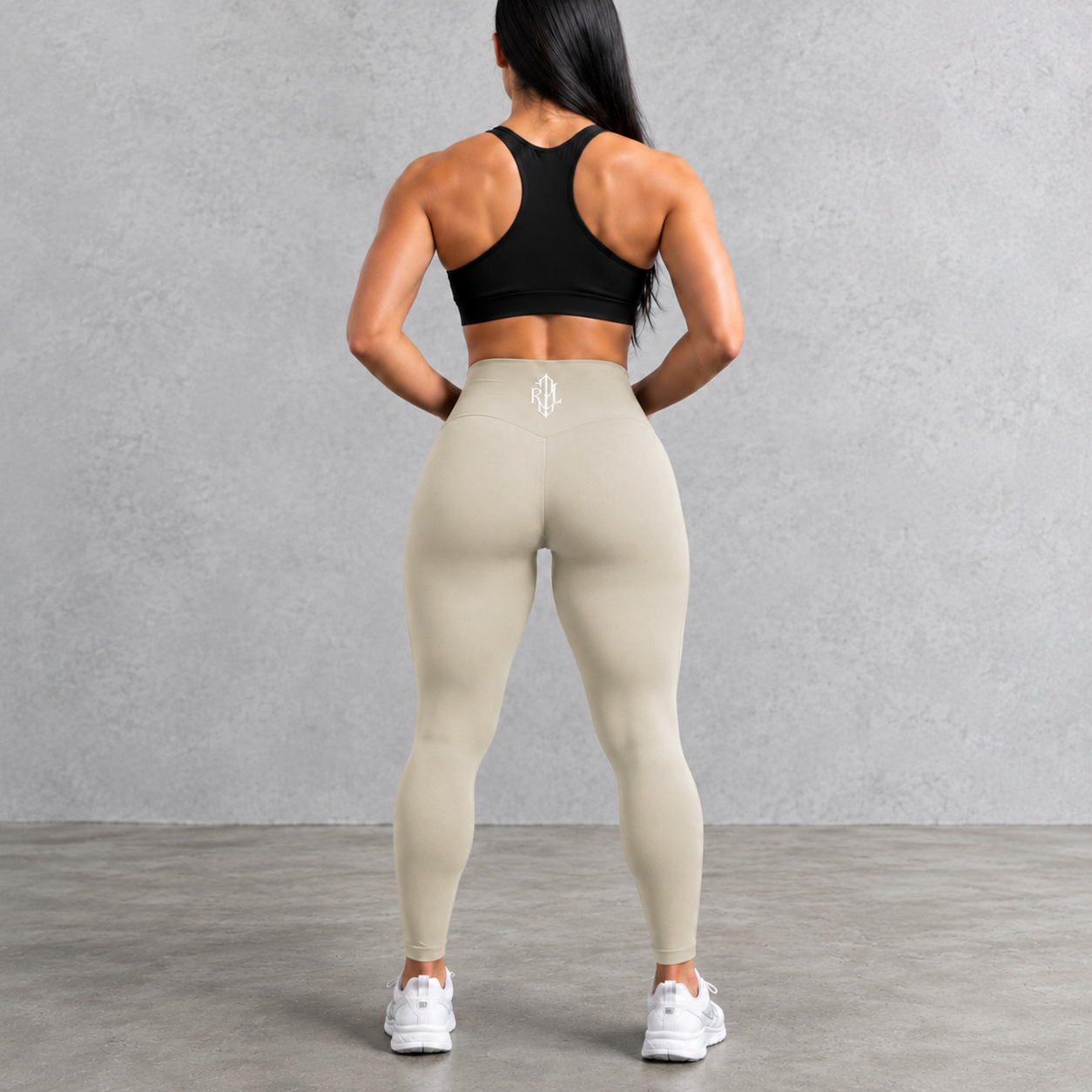 Rear view of a woman wearing beige high-waisted leggings and a black sports bra against a gray concrete wall and floor.