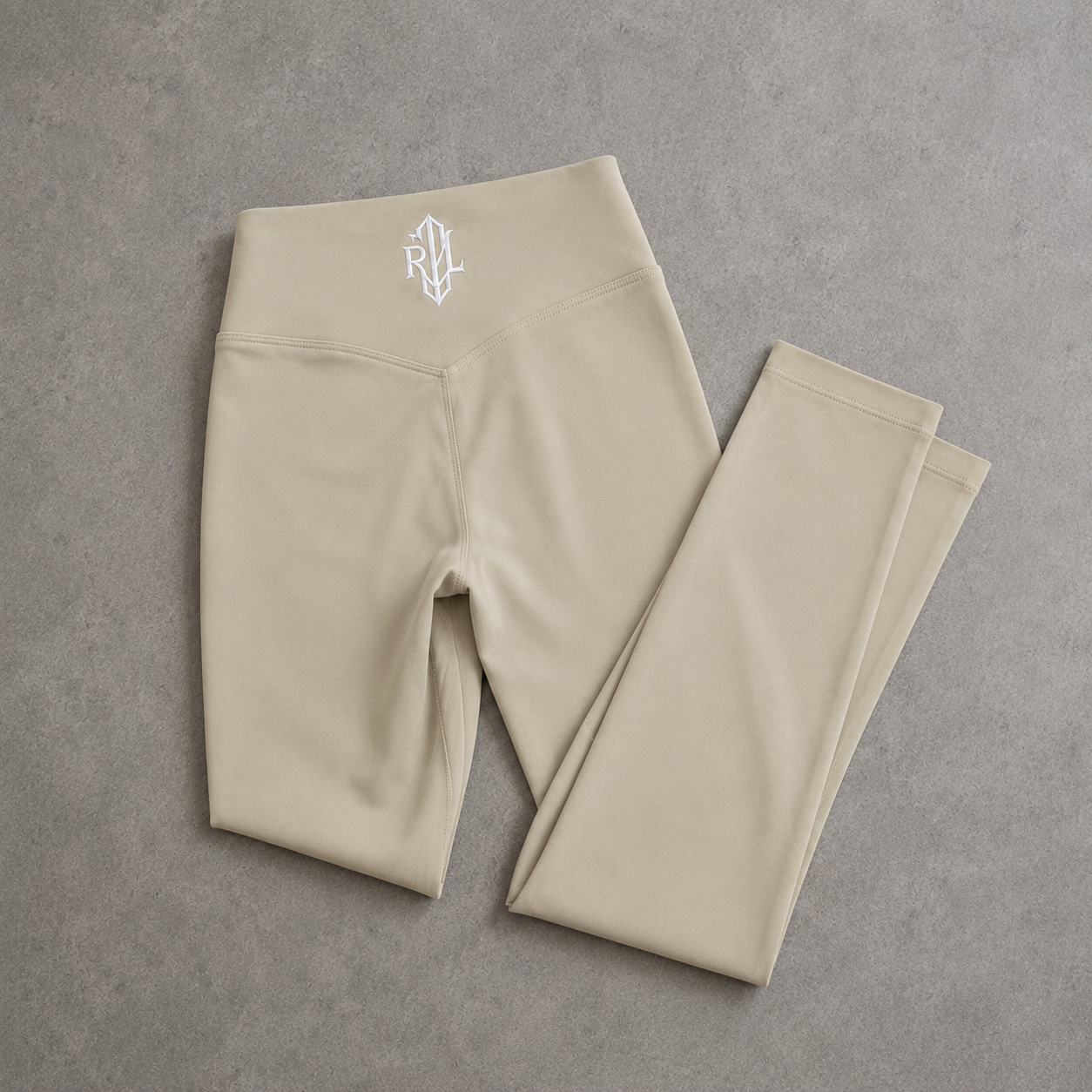 Beige high-waisted leggings folded neatly on a textured gray surface, featuring a small white monogram logo on the waistband.