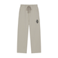 A pair of light beige drawstring sweatpants with a small colorful graphic patch on the left hip, displayed against a solid white background.