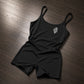 A black sleeveless romper with thin shoulder straps and a small white stylized logo on the left chest, laid flat on a dark wood floor.