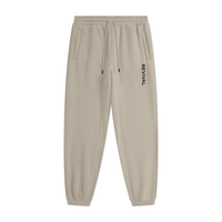 Beige tapered jogger pants with an elastic drawstring waistband, side pockets, elastic cuffs, and vertical black REVIVAL lettering on the upper thigh.

