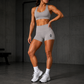 A woman modeling a light gray athletic set consisting of a sports bra and high-waisted shorts with a small black logo on the left leg, standing in a dark gym environment.