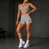A woman modeling a light gray athletic set consisting of a sports bra and high-waisted shorts with a small black logo on the left leg, standing in a dark gym environment.